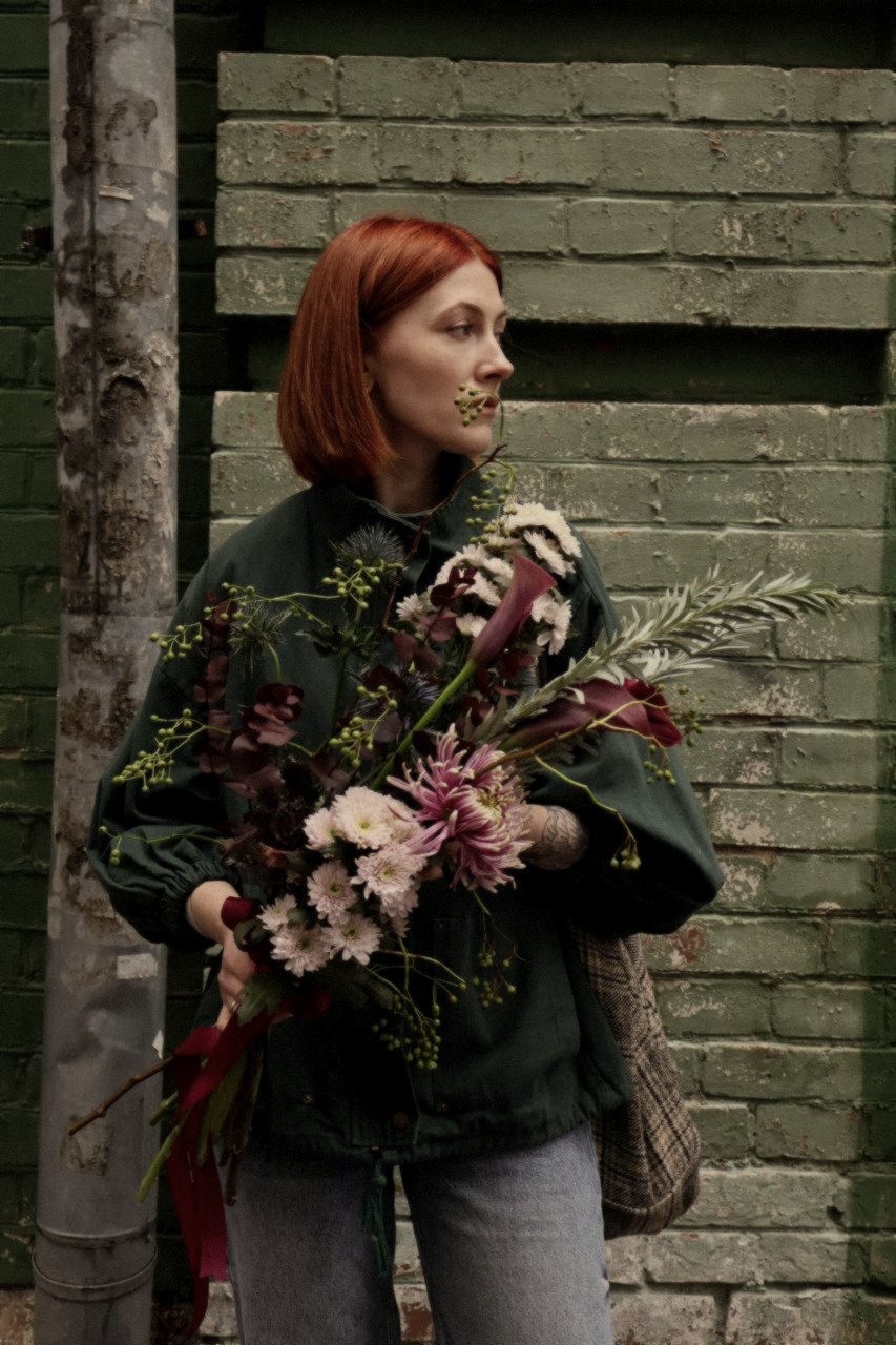 Red hair, a work jacket, and a bouquet against a peeling green wall