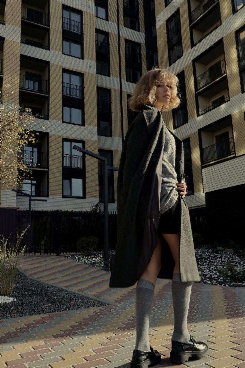 A woman in a long charcoal coat and grey socks stands before a new building
