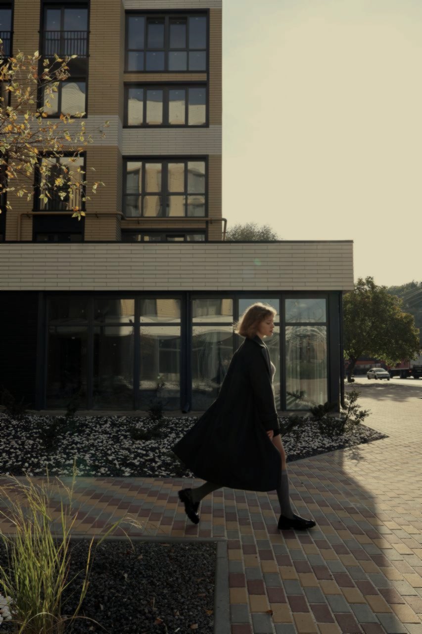 A woman walks in a long charcoal coat, grey socks left visible, loafers heavier than the dress